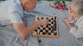 Above view of two people of mature age lying down on stomach on grey rug and playing chess. Grey-haired husband and wife spending great time outdoors. Concept of active life on retirement. - Powered by Shutterstock - Get 15% off with code: PIKWIZARD15