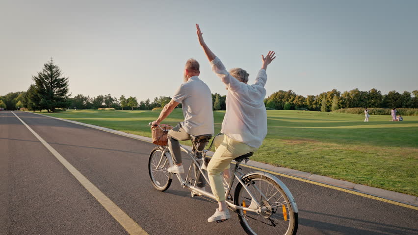 Back view of elder couple traveling on riding path with double bicycle. Senior man and woman engaging in healthy lifestyle while moving near golf course. Concept of retirement and transport.