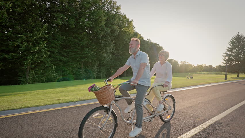 Side view of lovely grey haired wife and husband using eco transport while breathing fresh air and healing their bodies. Happy caucasian married couple relaxing and riding double bicycle together.