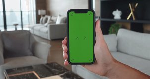 Male hold mobile phone in hand with chroma key green screen at living room. Close up mobile phone with green chroma key mock up screen in vertical mode at home background. Using phone, watching video