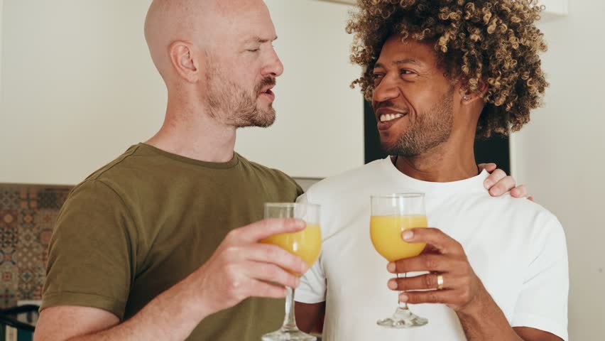 Black man with curly hair and a caucasian man as a Happy gay couple drinking orange juice and having a healthy breakfast at kitchen, while smiling and talking to each other.