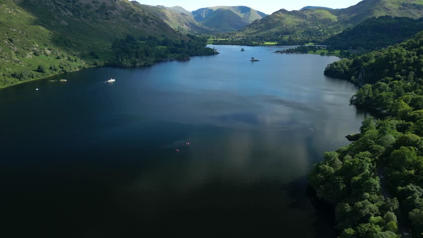 Dark lake surrounded by wooded mountains with flight towards white ferry on bright summer day. Ullswater, English Lake District, Cumbria, UK.