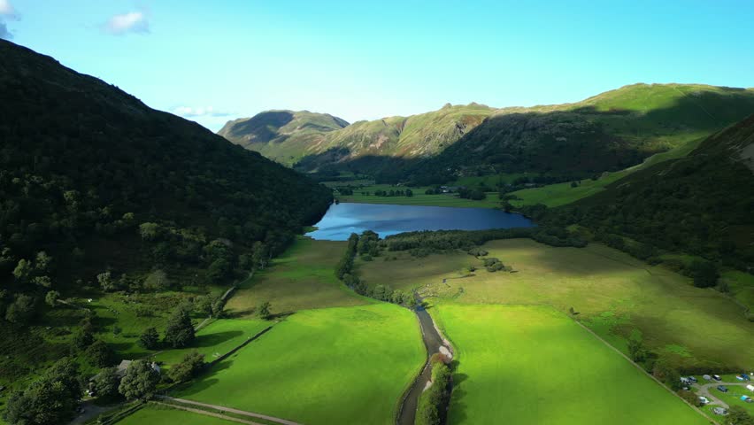 Sunlit pools moving across lush green patchwork fields on valley floor with wooded mountainsides and distant lake Brotherswater on summer day. English Lake District, Cumbria, UK.