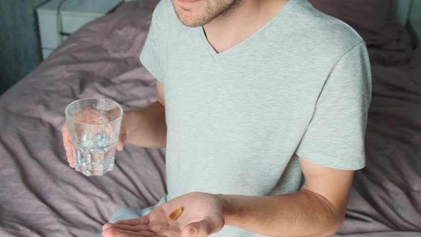 Dietary supplements. Man drinking omega 3 capsule with glass of water while sitting on the bed. Morning dose of vitamin D. Fish oil in pills