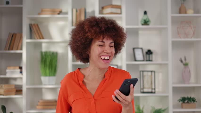 Multi ethnic female employee expressing happiness while looking on smartphone screen. Emotional dark haired lady in orange blouse reacting on good news with sincere emotions at workplace Royalty-Free Stock Footage #1108314105