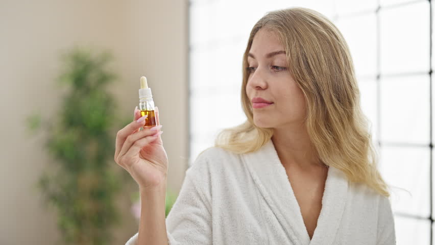 Young blonde woman wearing bathrobe holding face oil bottle at home