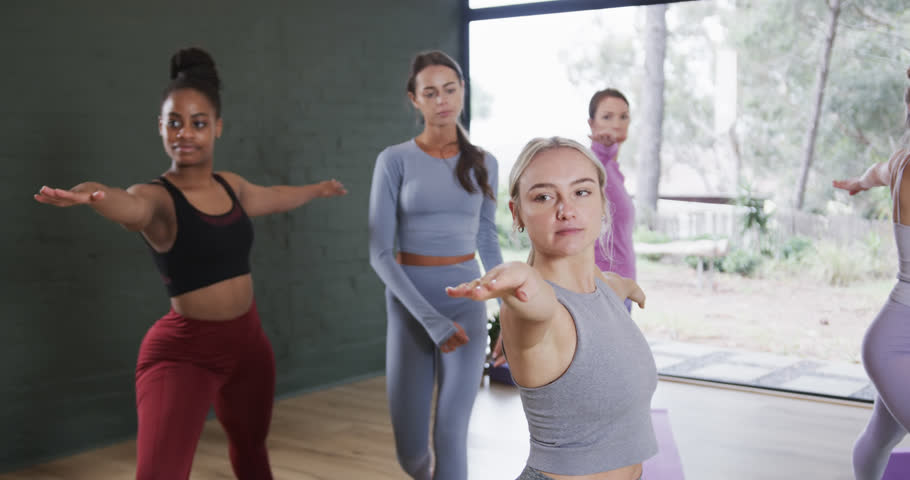 Diverse female instructor helping women in practicing warrior 2 pose in yoga class. Unaltered, slow motion, yoga, exercise, fitness, zen, healthy lifestyle, smile and wellbeing. Royalty-Free Stock Footage #1108320859