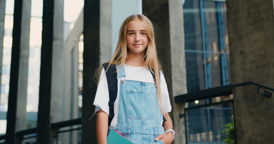 Pretty schoolgirl with loose long hair standing near school building, posing and looking at camera after classes, outdoors. Education concept