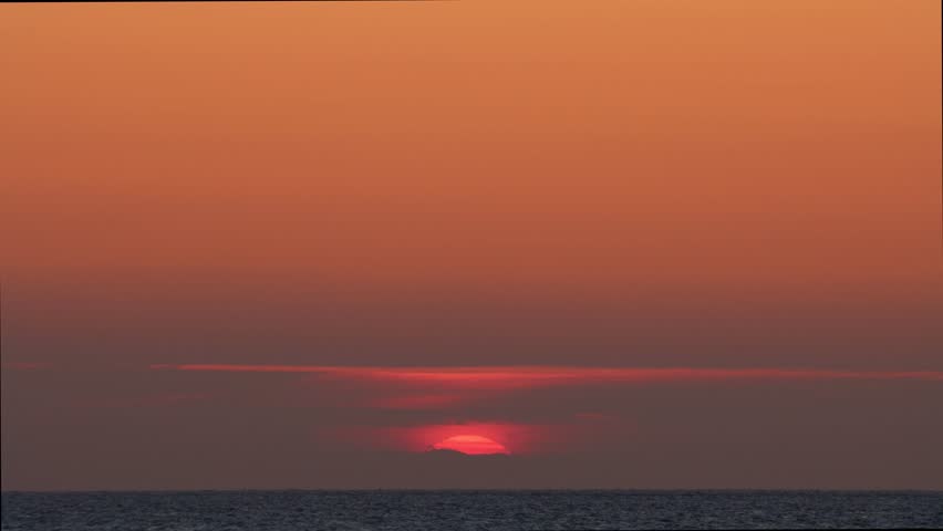 The sun setting over the horizon on the background of the calm blue sea. Wind turbines seen to the left of the sun.