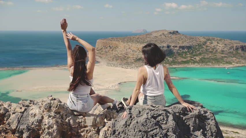 Two carefree beautiful young mix race asian women sitting back on the rocks observing blue lagoon sea coastline. Girls tourists have fun laugh together. Luxury resort. Best friends relationships