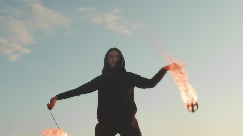 Medium shot of young professional male artist juggling with two burning staffs while performing fire show outdoors at sunset | Shutterstock HD Video #1108339549
