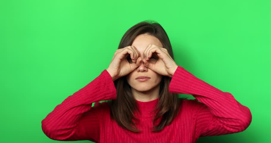 Amazed curious woman with brunette hair in sweater looking around through fingers imitating binoculars and point at something surprised. Indoor studio shot isolated on green background. Royalty-Free Stock Footage #1108345679