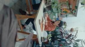 Female owner of flower shop working on laptop and drinking tea from cup, sitting at desk in cozy office decorated with green plants. Vertical clip, full length