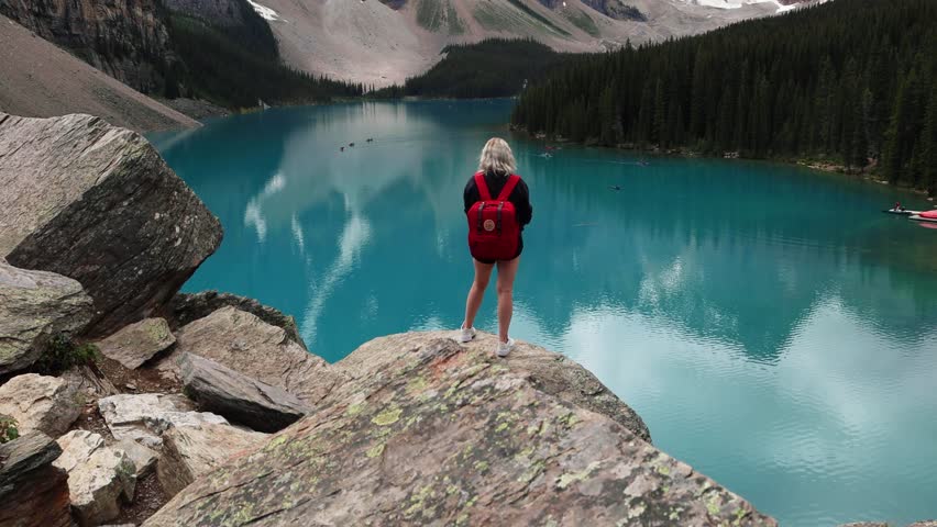 Looking our over the bright blue glacial water of Moraine Lake with tall jagged mountains and dense pine forests, Banff National Park, Alberta, Canada. Traveller with red backpack. Royalty-Free Stock Footage #1108347429