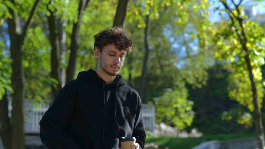Happy guy enjoying coffee while walking in the city park, talking on the mobile phone. Lifestyle. Concept of autumn walks. Slow motion