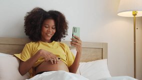 Smiling Black African American Pregnant woman holding showing ultrasound scan photos. Expecting mother having video call with family on smartphone at bed. - Powered by Shutterstock - Get 15% off with code: PIKWIZARD15