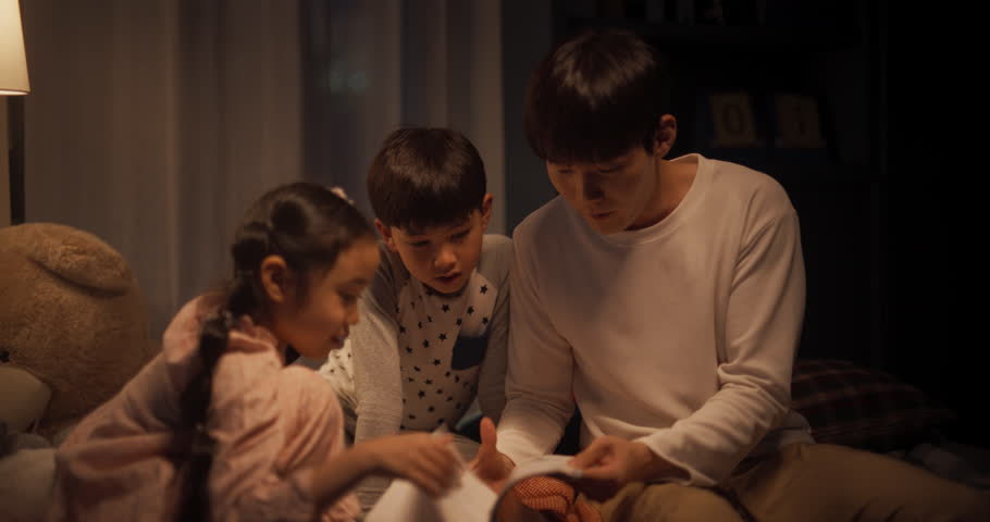 Korean Father Reading a Fairytale to His Lovely Cute Kids in Bed Before Going to Sleep. Young Handsome Dad Caring for his Little Children, Reading Stories from a Book in the Evening at Home