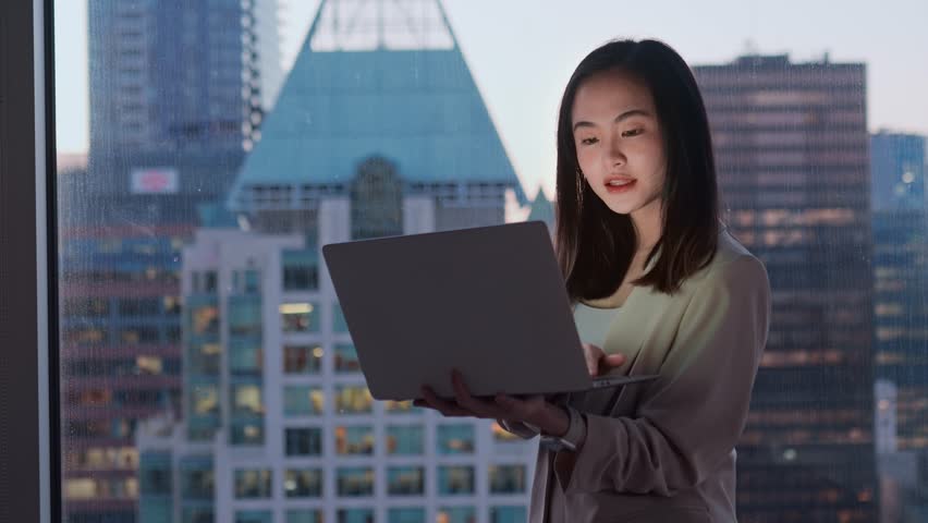 Young confident busy Asian business woman executive holding laptop in dark office. Professional businesswoman manager using computer standing at window with big city evening view. Royalty-Free Stock Footage #1108358489