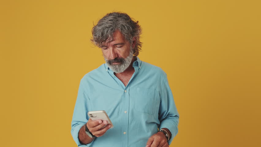 Elderly grey-haired bearded man wears a blue shirt, looks enthusiastically at a mobile phone, points to the phone while looking at the camera, isolated on an orange background in the studio - Powered by Shutterstock - Get 15% off with code: PIKWIZARD15