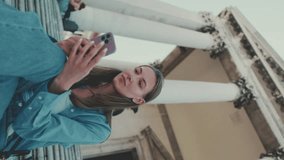Vertical video, Cute girl with long brown hair, wearing blue shirt, uses mobile phone while sitting on steps in Venice