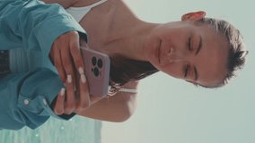 Vertical video, Beautiful girl with long brown hair, dressed in blue shirt, sits on embankment in Venice and uses mobile phone, boats and speedboats on background