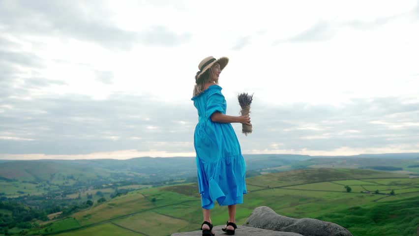 asian woman in a blue dress and a hat with a lavender bouquet in her hands on a top of mountain at summer sunset	