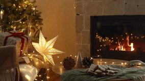 Cozy christmas eve in festive decorated living room. Stylish gifts and gingerbread cookies on table against christmas tree with lights and burning fireplace. Winter hygge footage
