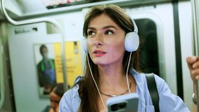 Female chatting online in mobile phone messenger watch video in metro. Young girl listening music in headphones and hold smartphone in subway car travel underground use wireless internet connection.