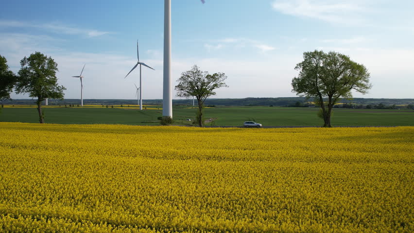 Aerial establishing shot showing driving cars on countryside road behind golden rape field and Rotating wind turbines in background Royalty-Free Stock Footage #1108369487