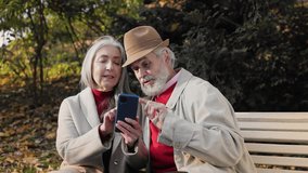 Happy old mature retired couple using smartphone for watching funny photos and video, mobile applications, social media, shopping online. Middle aged spouses enjoying time together in autumn park.