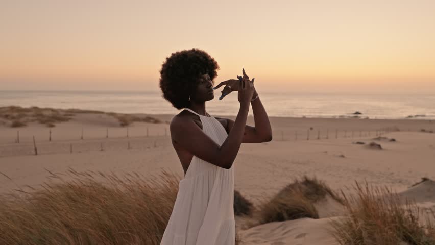 A beautiful black woman in a flowing white dress performs a conceptual dance on sandy beach dunes at sunset, connecting harmoniously with nature's rhythm and beauty Royalty-Free Stock Footage #1108373241