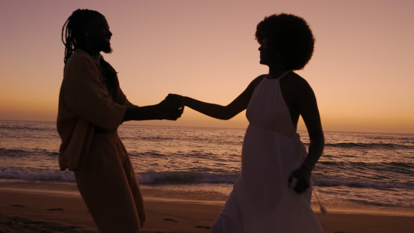 A black woman and African male, silhouetted against a mesmerizing sunset, walk on the beach, sharing smiles and flirtatious moments, radiating love and romance Royalty-Free Stock Footage #1108373245