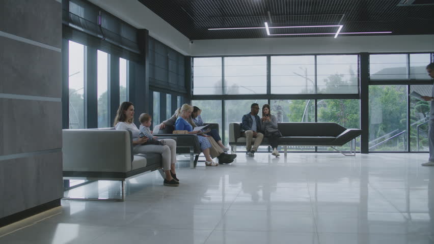 Diverse people sit on sofas in clinic lobby area, wait for appointment with doctor. Doctor speaks with elderly couple about medical test results. Waiting area in modern medical center. Healthcare. Royalty-Free Stock Footage #1108377873