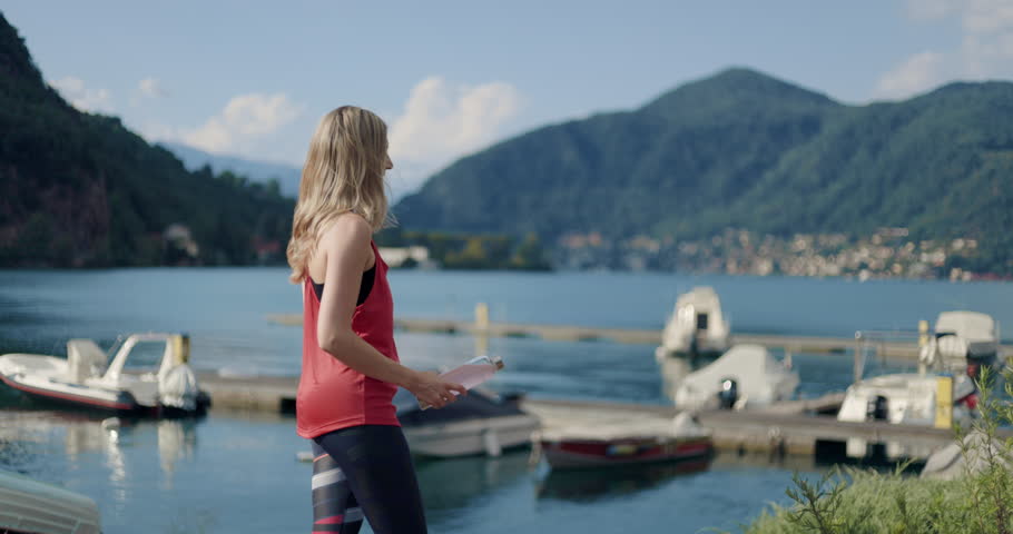 Woman runner takes a break on the lakeside to take a food supplement, vitamins, pills, medicine, energizing drink, proteins in a beautiful transparent bottle. Marina,small boats, alps, green mountains