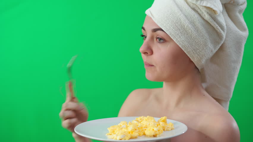 Young slim woman with omelette plate biting fork looking away and looking at camera shrugging shoulders. Portrait of Caucasian lady in hair towel posing with food at chromakey background