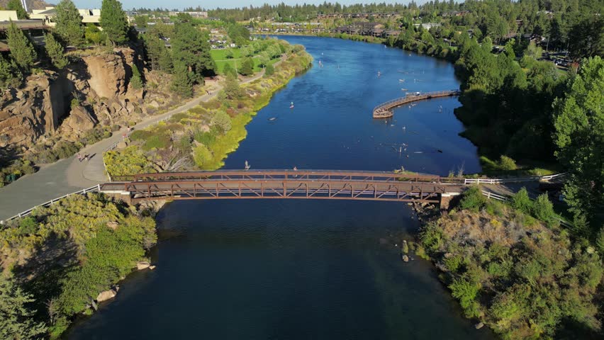 Aerial View Footbridge Over River in Bend Oregon USA. Approach 4k Drone with Paddle Board Athlete Family Activity near Local Town Park with Blue Water and Green Trees Athletic Health Wellness Spring
