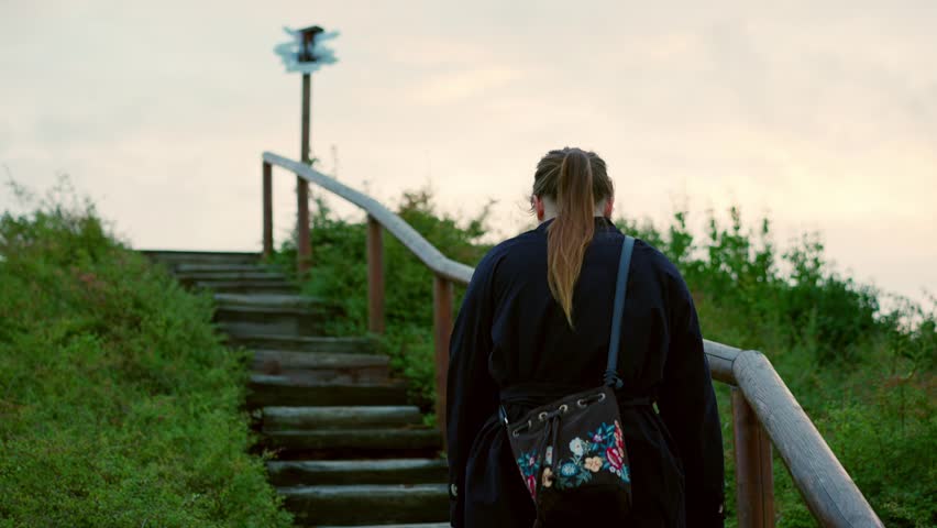 Girl in a long black coat with her hair in a ponytail is walking on the stairs in a nature park in autumn. Rear shot. Shot from behind