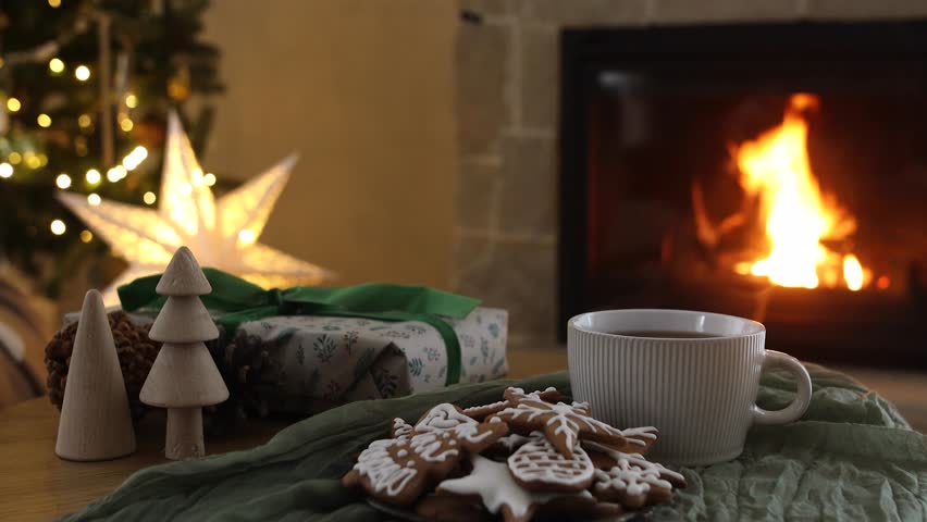 Cozy christmas eve. Stylish wrapped gift, gingerbread cookies and tea cup on table against festive christmas lights and burning fireplace. Winter hygge footage