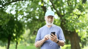 Senior active gray haired bearded man in headphones uses a smartphone while walking in urban city park. Mature old fitness male is chatting online, scrolling browsing social networks or choosing music - Powered by Shutterstock - Get 15% off with code: PIKWIZARD15