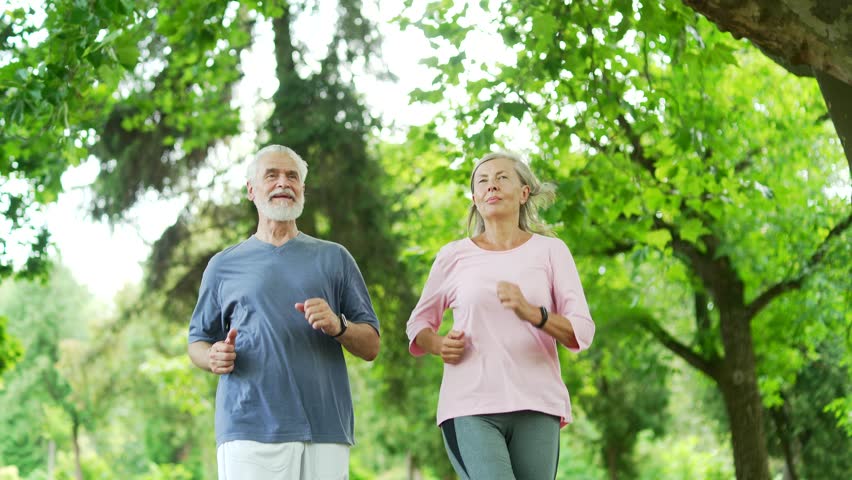 Active married old senior couple jogging in an urban city park. Sporty mature gray haired retired wife and husband enjoys running in nature in morning. Elderly runners during a joint workout in forest