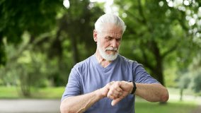 Close up. Senior active gray haired bearded runner looking smart watch standing in city park. Mature old male using fitness tracker and checking result. Elderly sport man on training workout in nature - Powered by Shutterstock - Get 15% off with code: PIKWIZARD15