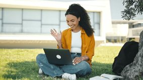 Online communication. Friendly pretty brazilian curly haired girl, successful student or freelancer, sit outdoors on a grass, talking on a video conference by laptop with friends or colleagues, smile