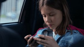 Female teen sitting on back seat in the car while holding mobile phone and playing game. Travel, technology, childhood concept. Real time