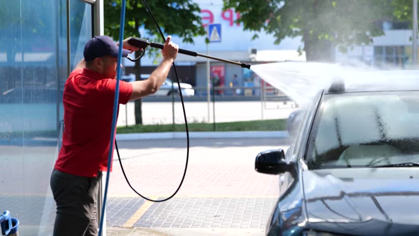 Young man in cap using a hose with pressurized water while washing his car at self-service car wash station. Self service, car wash concept. Real time Royalty-Free Stock Footage #1108389085
