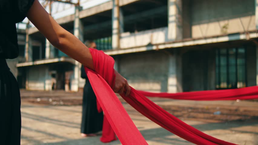 A close-up tracking shot of a dancer's arm in a black sleeve, firmly grasping a taut red fabric. The performance is set in an abandoned, sunlit industrial building, visible in the background.