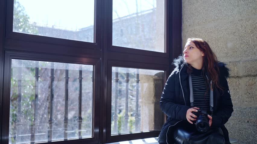 Young Latin woman tourist in warn clothes sitting near window and looking out with professional photo camera in Reina Sofia museum in Madrid, Spain during daytime