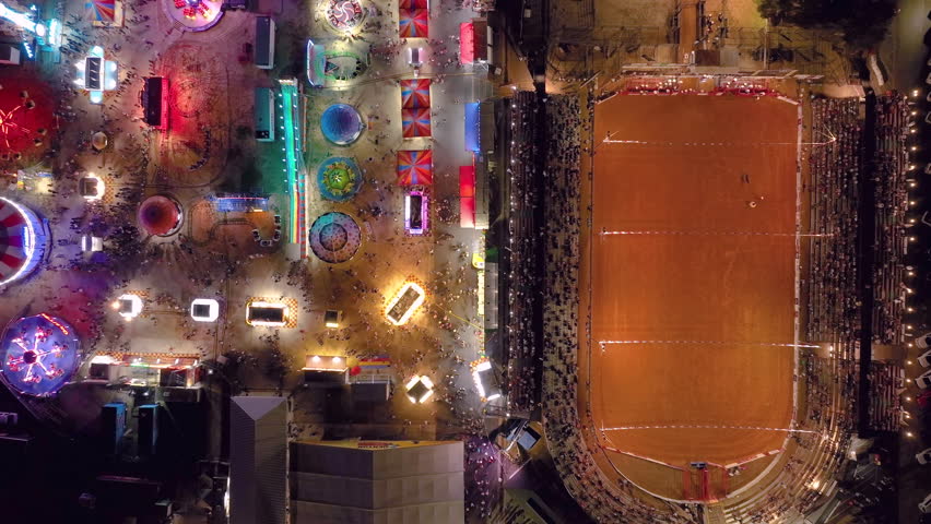 Aerial drone top down view of a carnival festival with colorful blinking neon lights on rides, attractions, and booths as the camera flies over a rodeo rink and the fairgrounds. | Shutterstock HD Video #1108393561