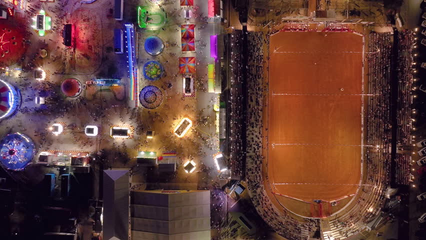 Aerial drone top down locked down view at night of a rodeo rink as a cow is let out of the pen and a cowboy chases it around the arena as a crowd watches with a colorful fair is seen on the left. Royalty-Free Stock Footage #1108393563