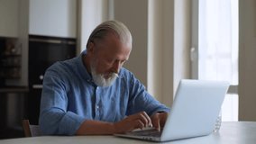 Senior aged bearded man talks via laptop at home. Communication, online meeting and businessman working on computer in living room with fresh water. Useful habits and healthy lifestyle, drinks water.