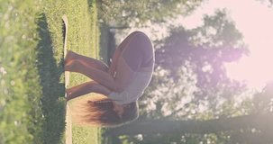 Yoga, woman gets into asana bakasana pose, standing on hands, balance. Side view, city park, lawn, green trees, morning sunlight, vertical video